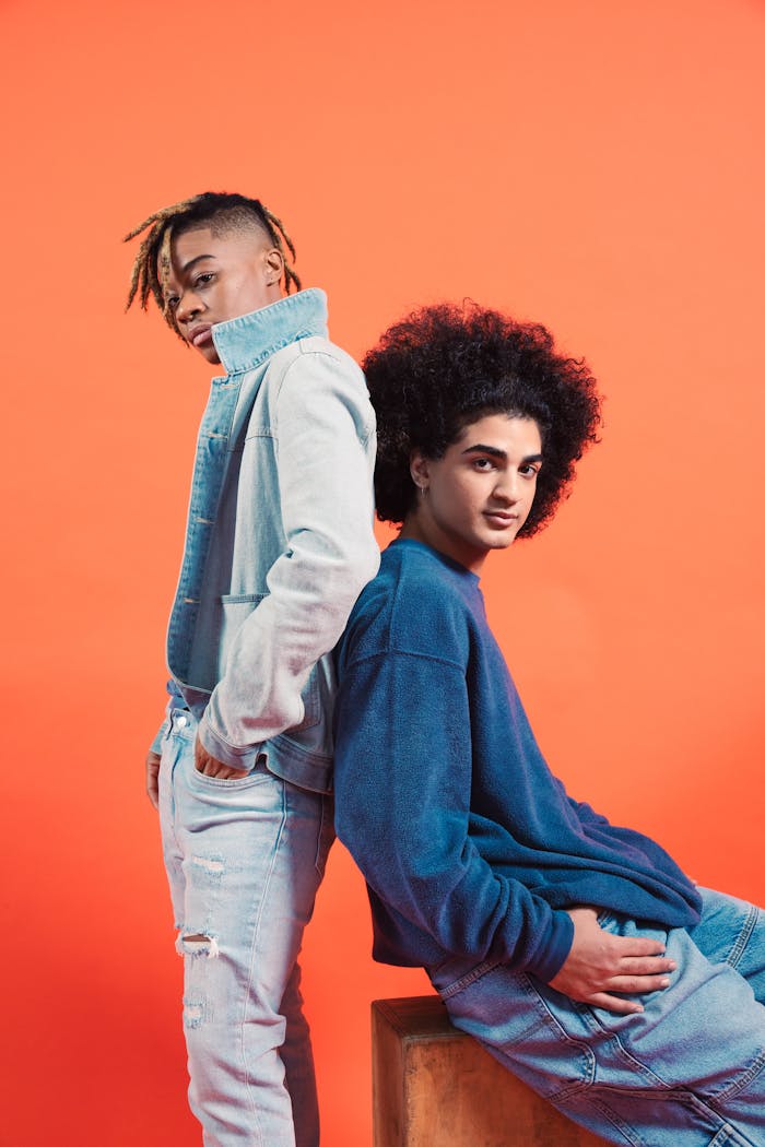 Two men pose back to back in denim fashions against an orange background.