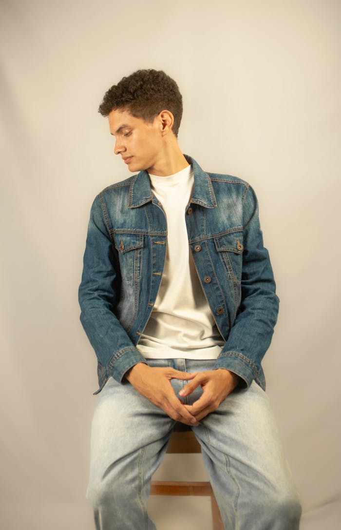 Young man wearing denim jacket and jeans, sitting on a stool indoors.