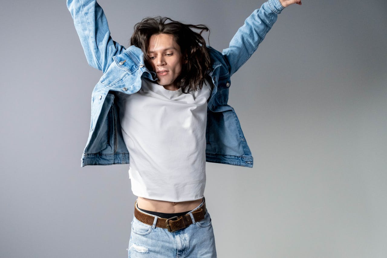 get-in-touch Young man with long hair jumps energetically wearing a stylish denim jacket in a studio setting.