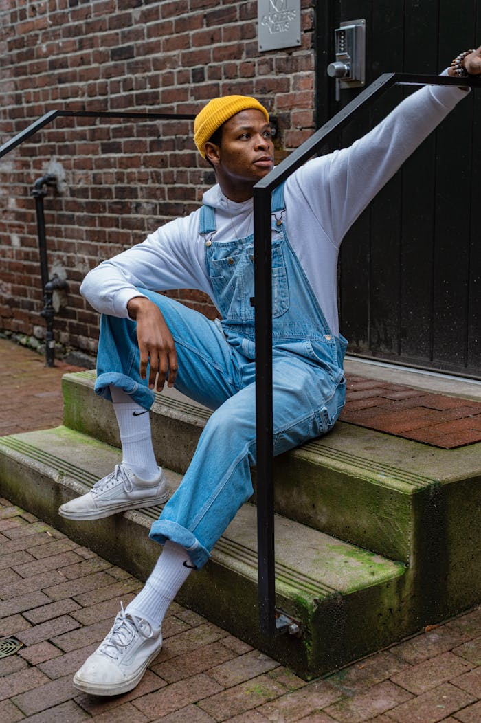 A stylish man poses outdoors in denim overalls and a beanie, showcasing urban fashion.